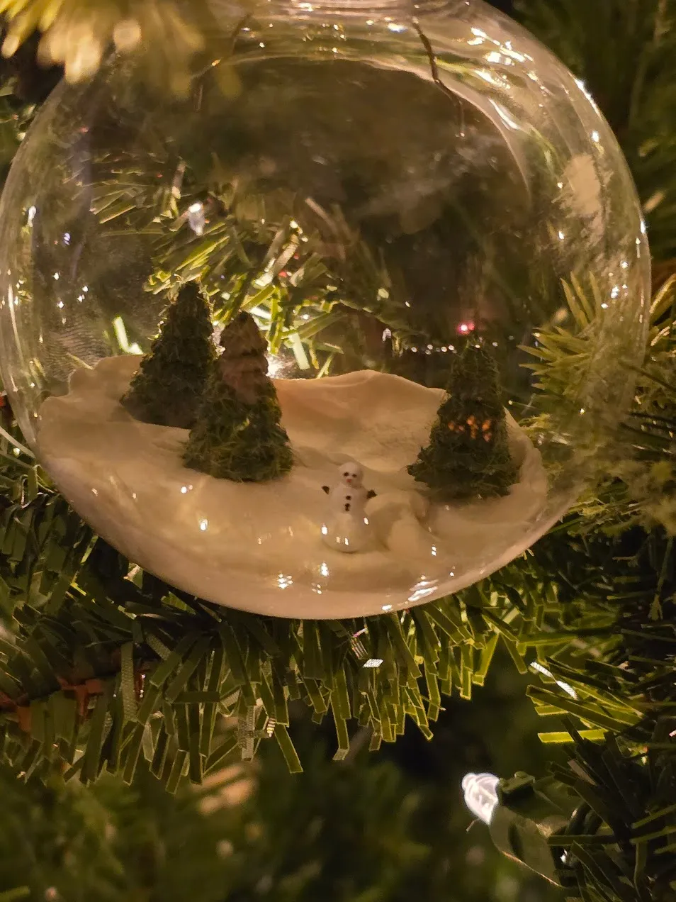 Clear glass ornament globe containing a tiny clay snow scene with three trees and a snowman